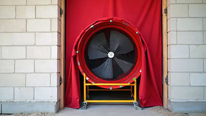 Large fan on red fabric in doorway of brick building. Equipment tests house airtightness. Energy audit checks air leaks. Safety standards inspection for building envelope. Saves money on heating